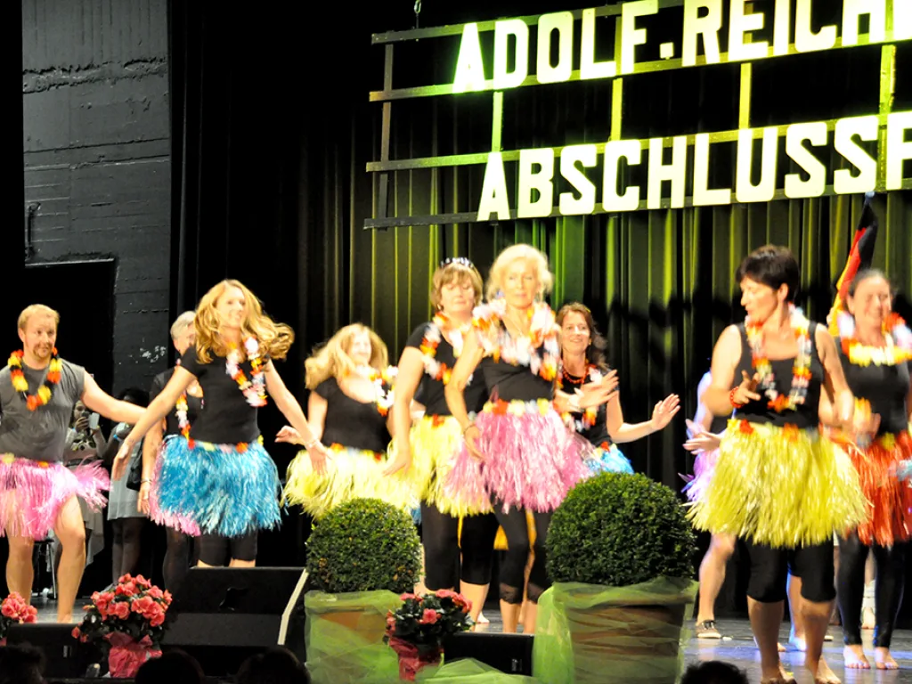 Tanzgruppe in bunten Röcken tanzt vor der beleuchteten Schriftwand.